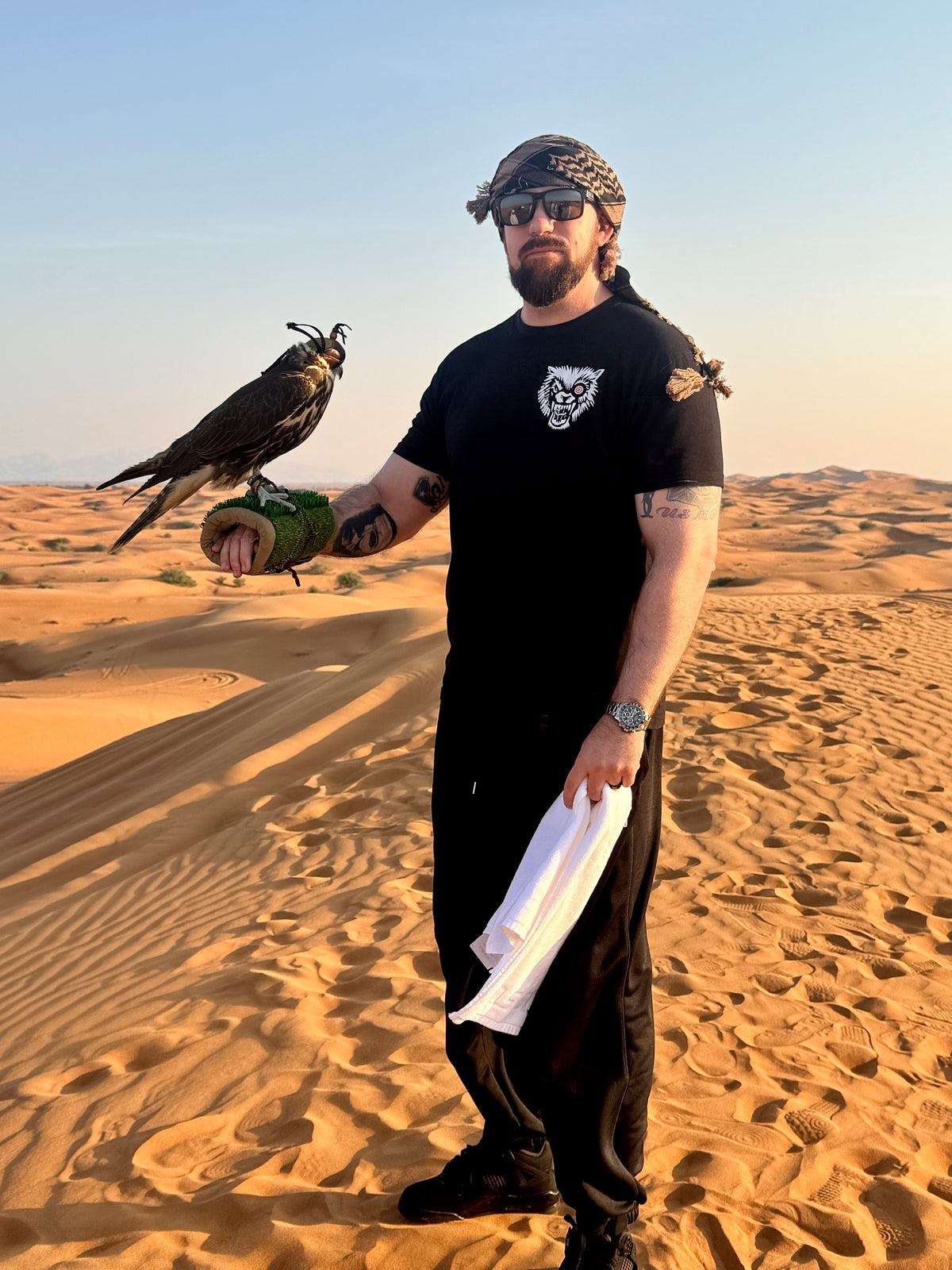 Man in desert wearing black T-shirt with bold white wolf logo, holding a falcon on his gloved hand.