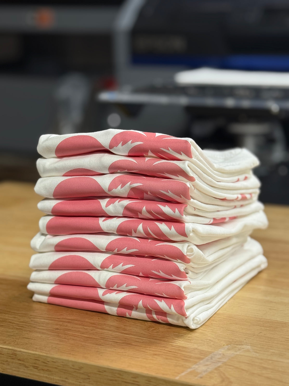 Folded cream T-shirts with pink logo print stacked on a wooden table, blurred workspace in the background.
